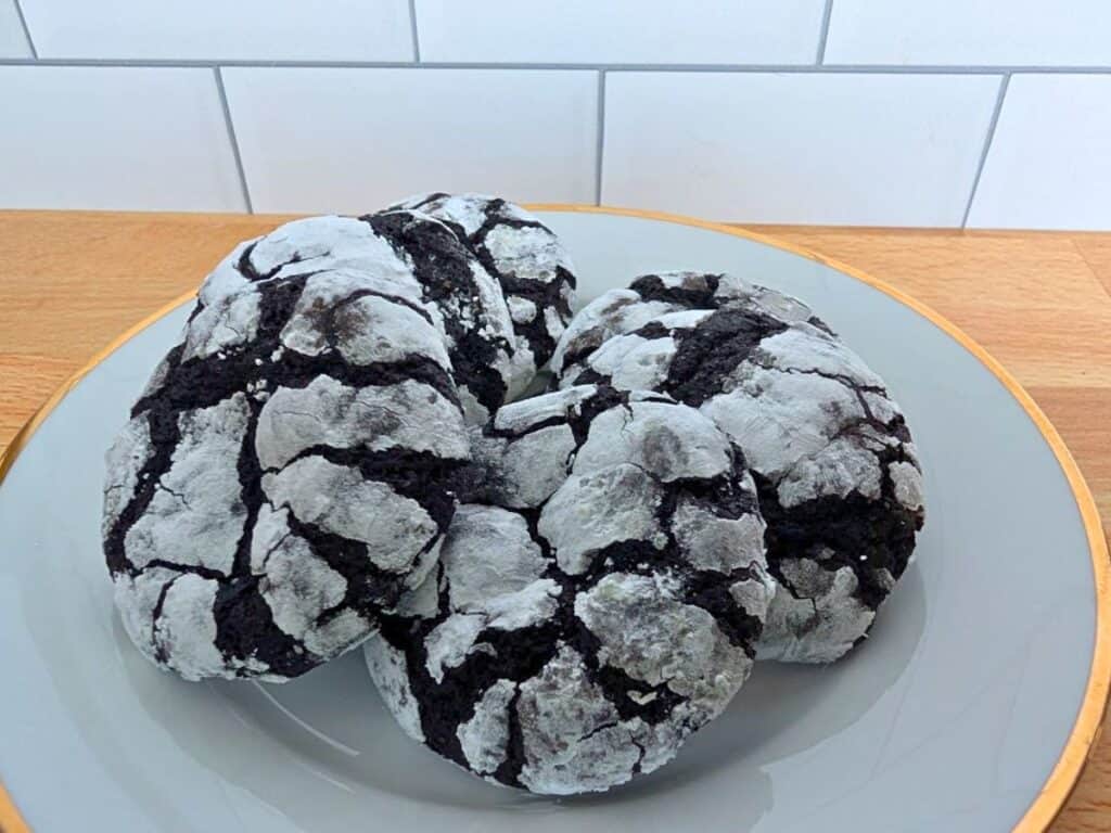 Four chocolate crinkle cookies coated in powdered sugar are arranged on a gray plate, set on a wooden surface with a white tile background.