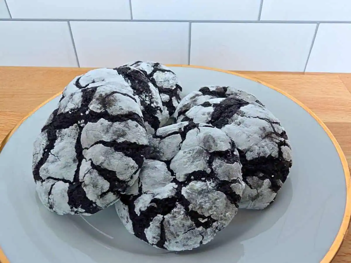 Four chocolate crinkle cookies coated in powdered sugar are arranged on a gray plate, set on a wooden surface with a white tile background.