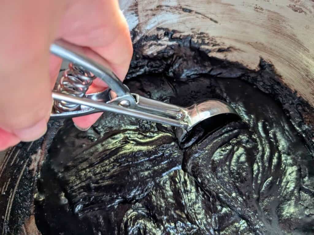 A hand uses a metal scoop to lift dark chocolate batter from a mixing bowl.