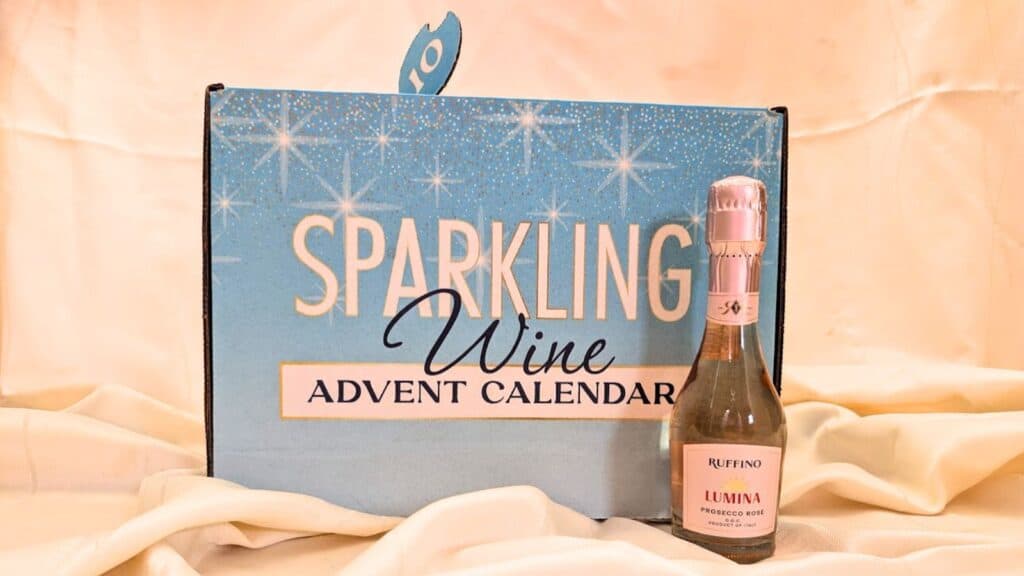 A blue box labeled "Sparkling Wine Advent Calendar" sits beside a small bottle of Ruffino Lumina Prosecco, making it a festive addition to Advent calendars, all displayed on a cream-colored fabric background.