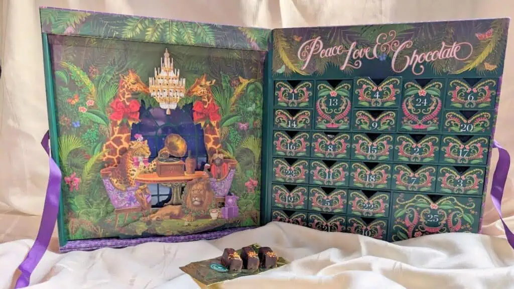 An open advent calendar decorated with giraffes, a chandelier, and tropical plants features three chocolates placed on a gold tray in front—a whimsical twist on traditional Advent calendars.
