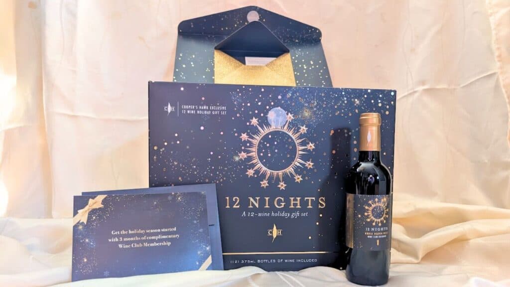 A festive "12 Nights" holiday wine gift set inspired by Advent calendars, featuring a decorated box, bottle of wine, and a card for a complimentary wine club membership—beautifully displayed against a light fabric background.
