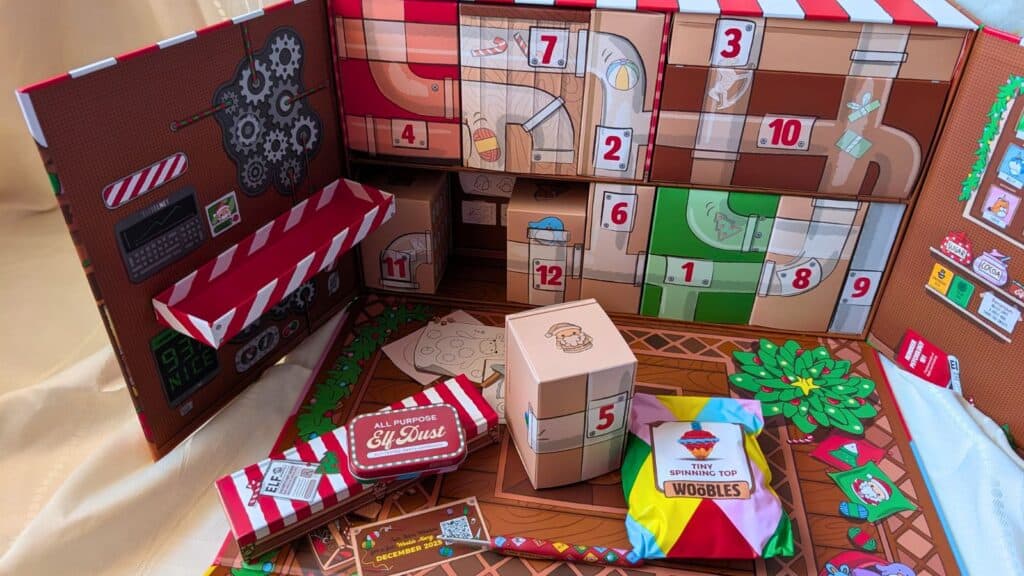 Open advent calendar with numbered compartments, some open to reveal small boxes and colorful packages on a decorated background featuring candy and mechanical illustrations.