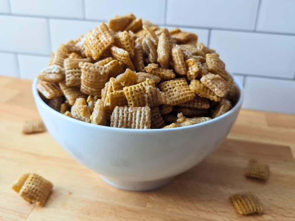 A white bowl filled with Chex cereal sits on a wooden surface with a white tiled background.