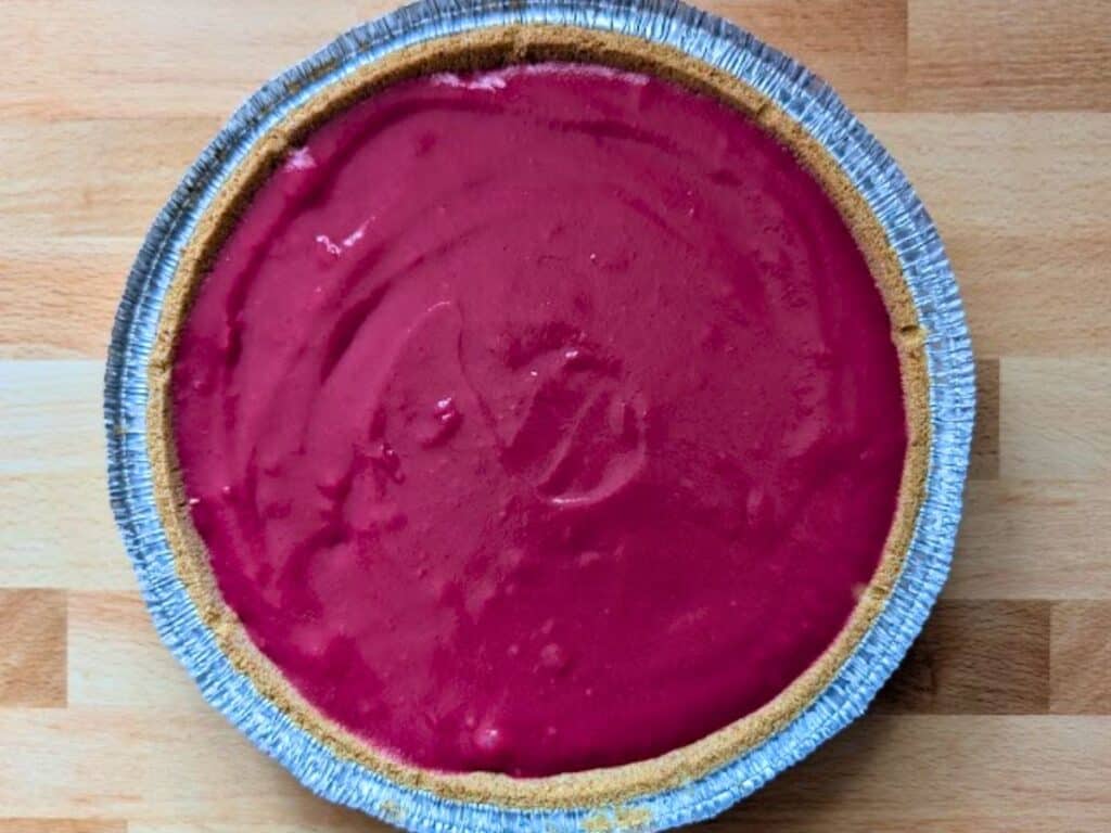 A graham cracker pie crust filled with a smooth, bright magenta filling, in a disposable aluminum pie tin, on a wooden surface.