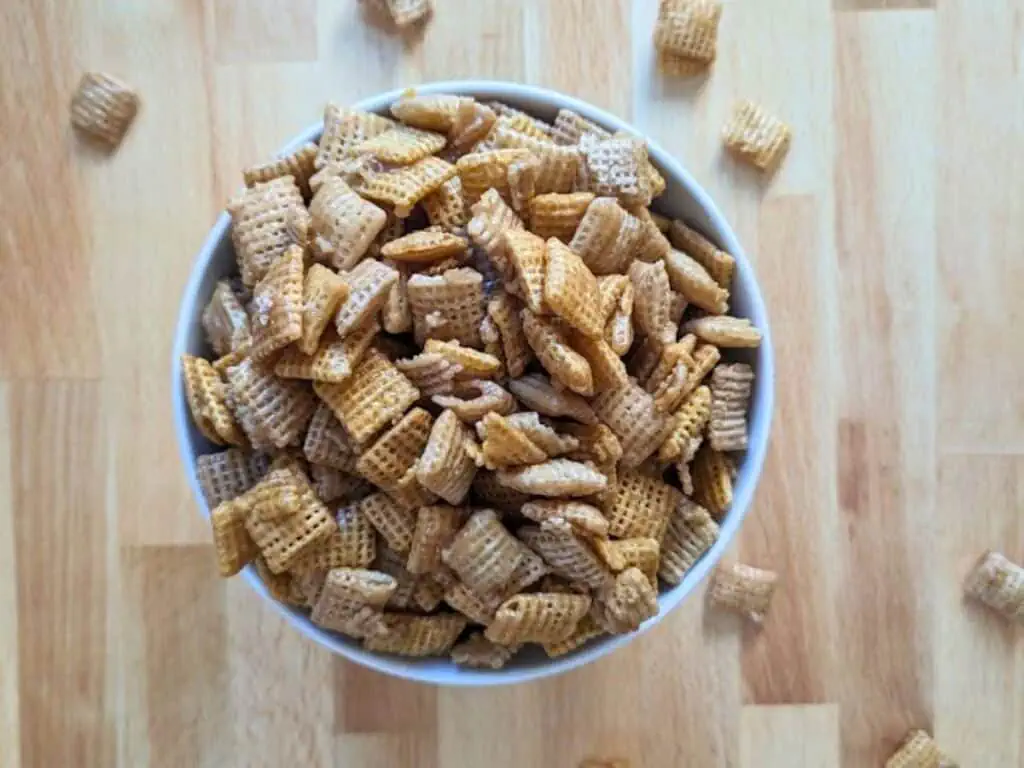 A white bowl filled with squares of Chex cereal sits on a light wooden surface, with a few cereal pieces scattered around.