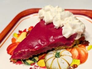 A slice of vibrant red pie with whipped cream on top, served on a decorative plate featuring autumn vegetables and pumpkins.