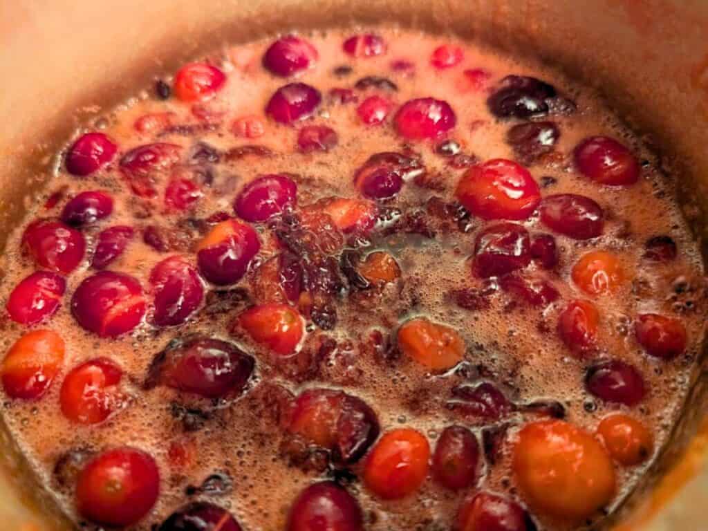 Whole cranberries simmer in a pot, creating a bubbling mixture with red and orange hues, likely part of a sauce or compote preparation.
