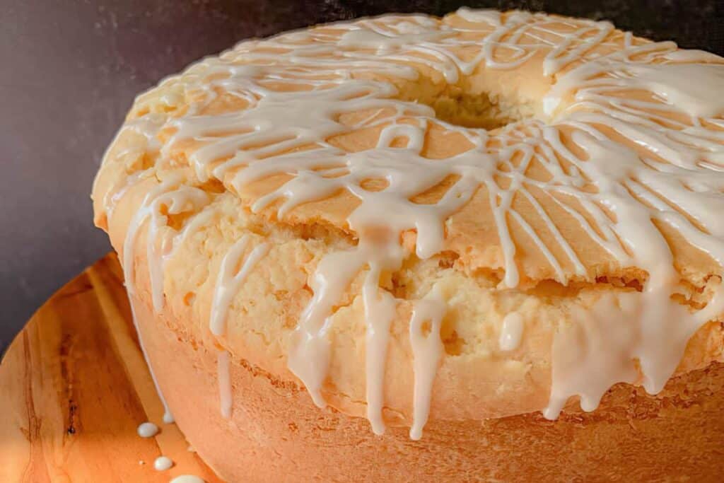 A round Bundt cake with a cracked top is covered in white icing glaze, sitting on a wooden surface.