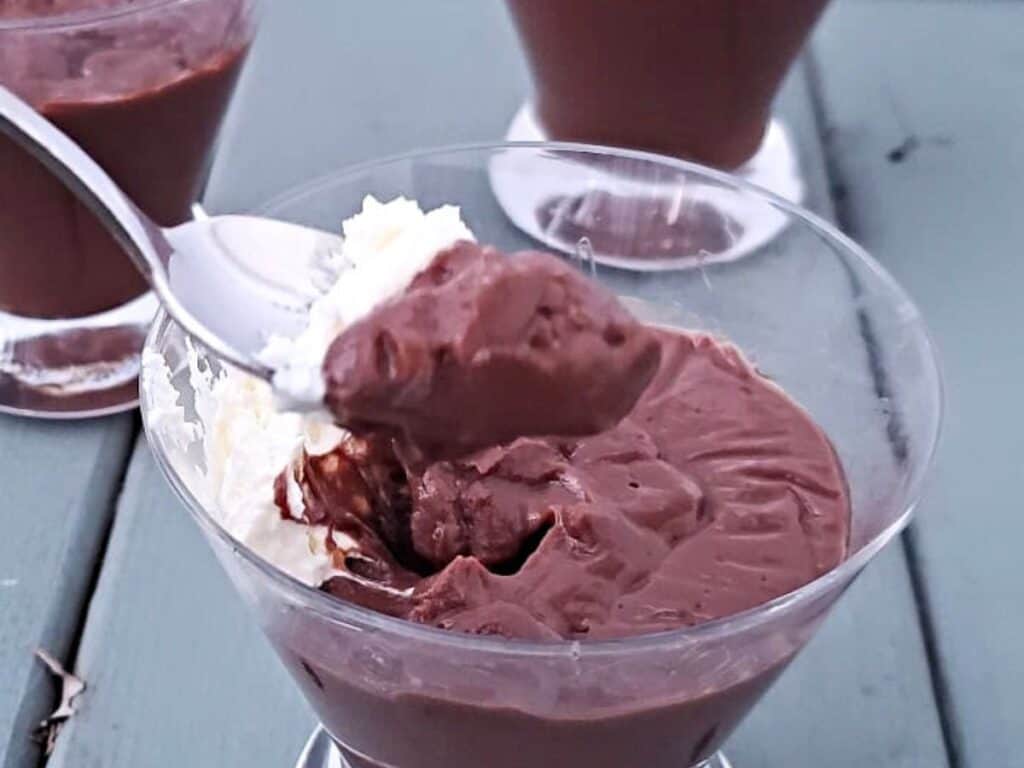 A close-up of a glass bowl filled with chocolate pudding and topped with whipped cream, with a spoon scooping some pudding. Two more bowls are in the background on a green surface.