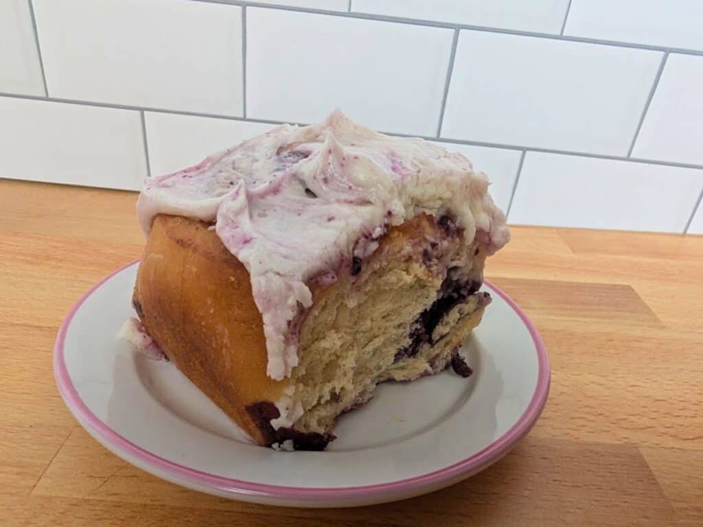 A single iced sweet roll with purple swirls, likely blueberry, sits on a small white plate with a pink rim on a wooden countertop in front of a white tiled wall.