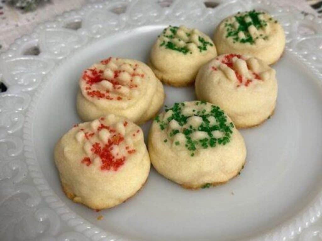 Six round shortbread cookies with red and green sprinkles arranged on a white decorative plate.