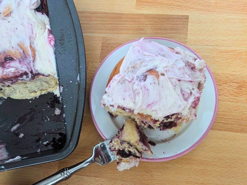 A frosted sweet roll with berry filling on a white plate, with a piece cut off by a fork. A baking tray with more rolls is partially visible on a wooden surface.