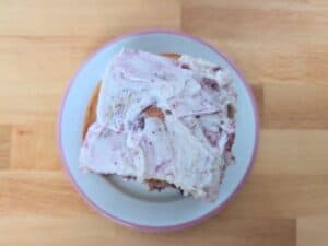 A cinnamon roll with berry-speckled frosting on a white plate, viewed from above on a wooden surface.