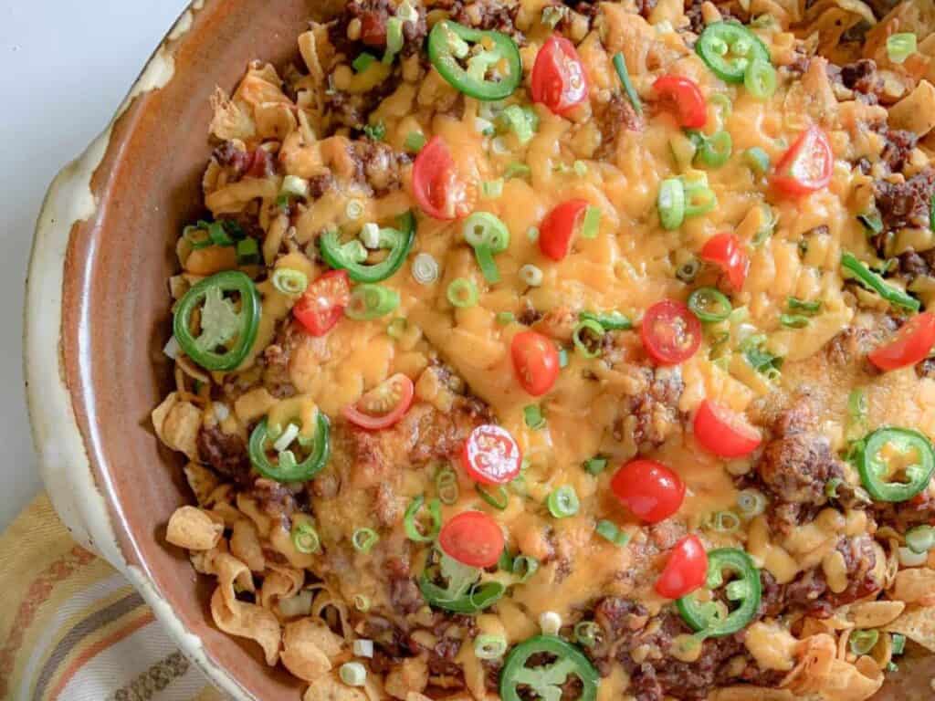 A dish with corn chips, melted cheese, seasoned ground beef, sliced jalape&ntilde;os, cherry tomatoes, and chopped green onions in a brown ceramic bowl.