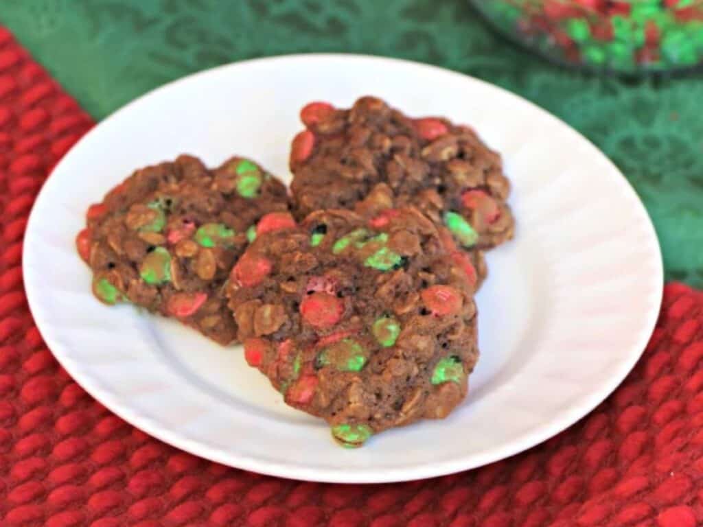 Three chocolate cookies with red and green candy pieces on a white plate, placed on a red textured surface.