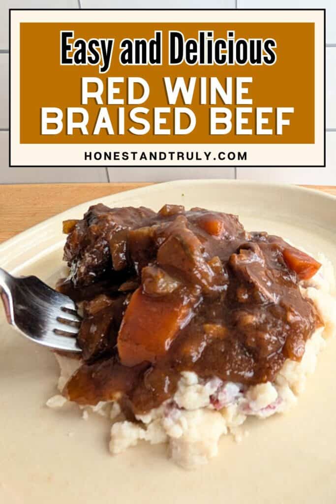 A serving of red wine braised beef with carrots and sauce sits on a bed of mashed potatoes; a fork is beside the food on a beige plate.