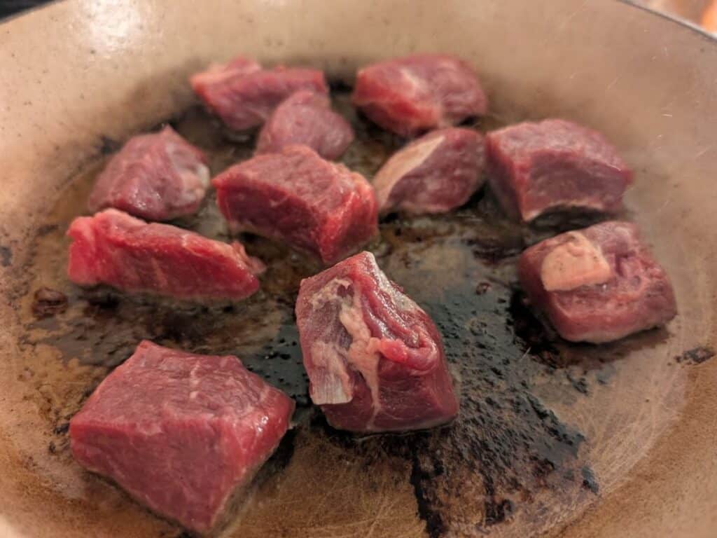 Chunks of raw beef are being seared in a pan, with some pieces beginning to brown while others remain red.
