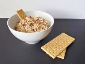A white bowl filled with a creamy dip and chocolate pieces, with a graham cracker inside. Two more graham crackers are placed beside the bowl on a dark surface.