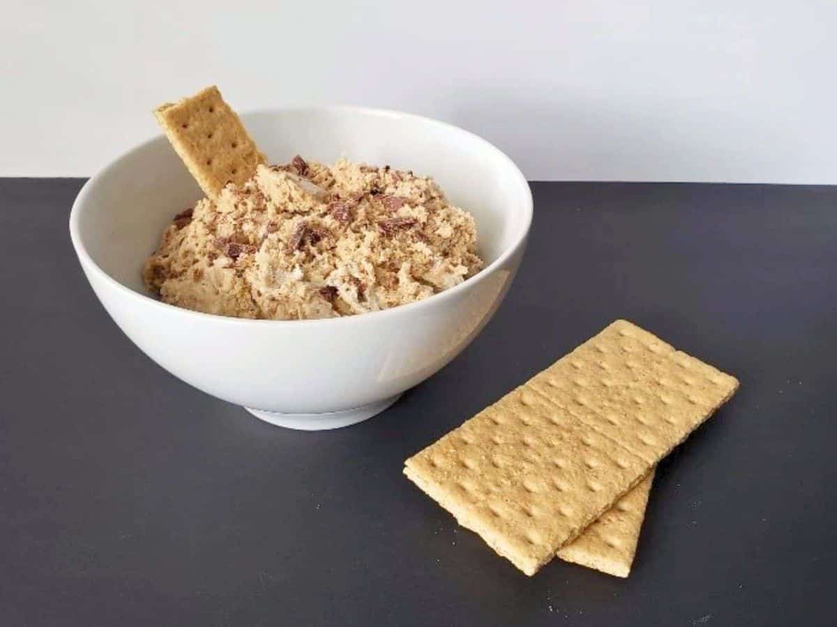 A white bowl filled with a creamy dip and chocolate pieces, with a graham cracker inside. Two more graham crackers are placed beside the bowl on a dark surface.