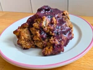 A large, irregularly shaped baked scone with a purple glaze sits on a white plate with a pink rim on a wooden surface.