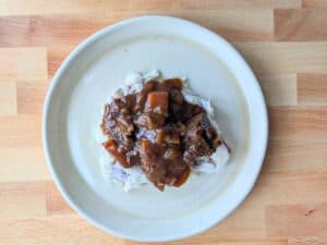 A plate of mashed potatoes topped with brown stew containing chunks of meat and vegetables, served on a light-colored wooden surface.