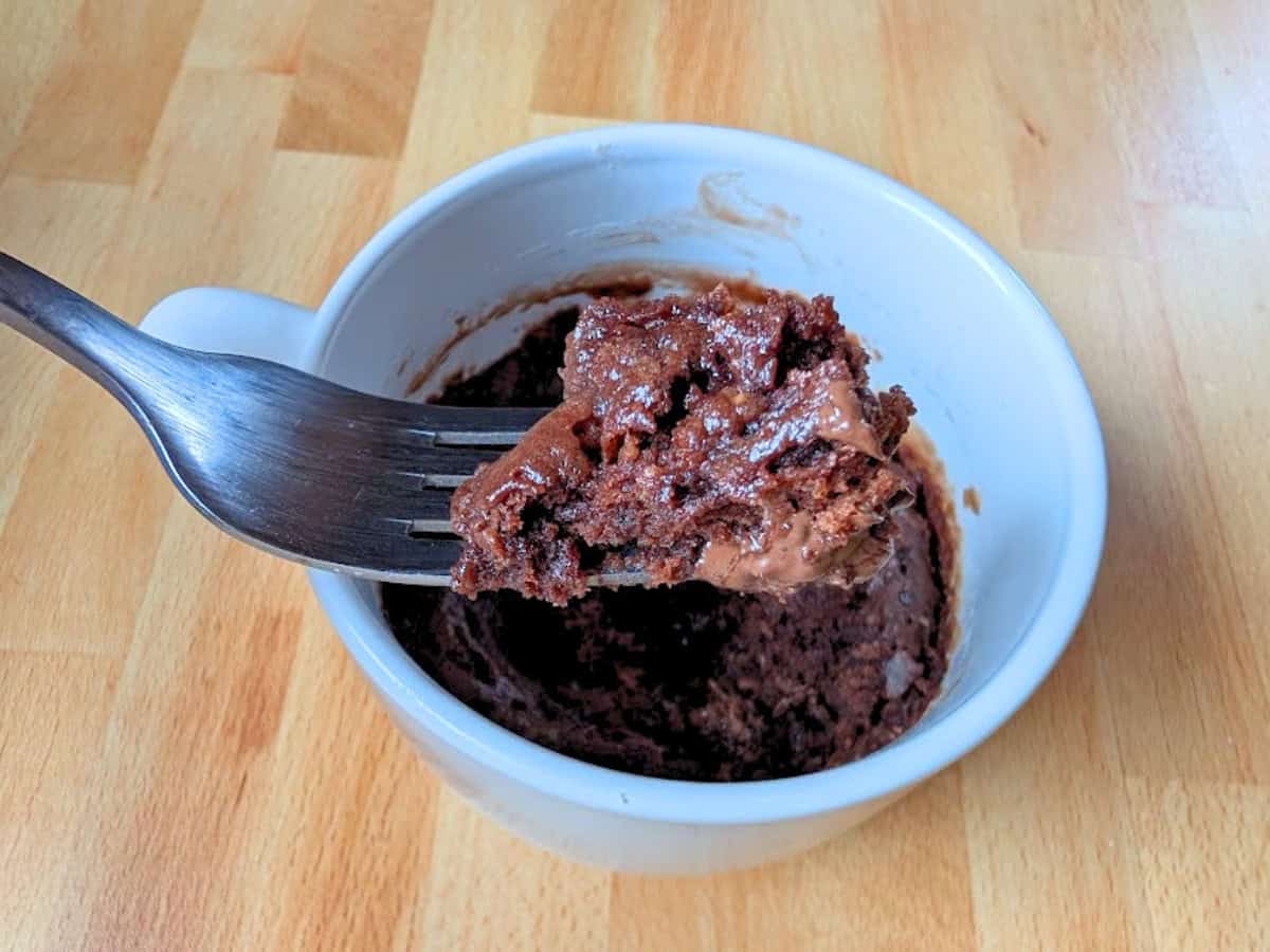 A fork holds a bite of chocolate mug cake above a white mug, sitting on a wooden surface.