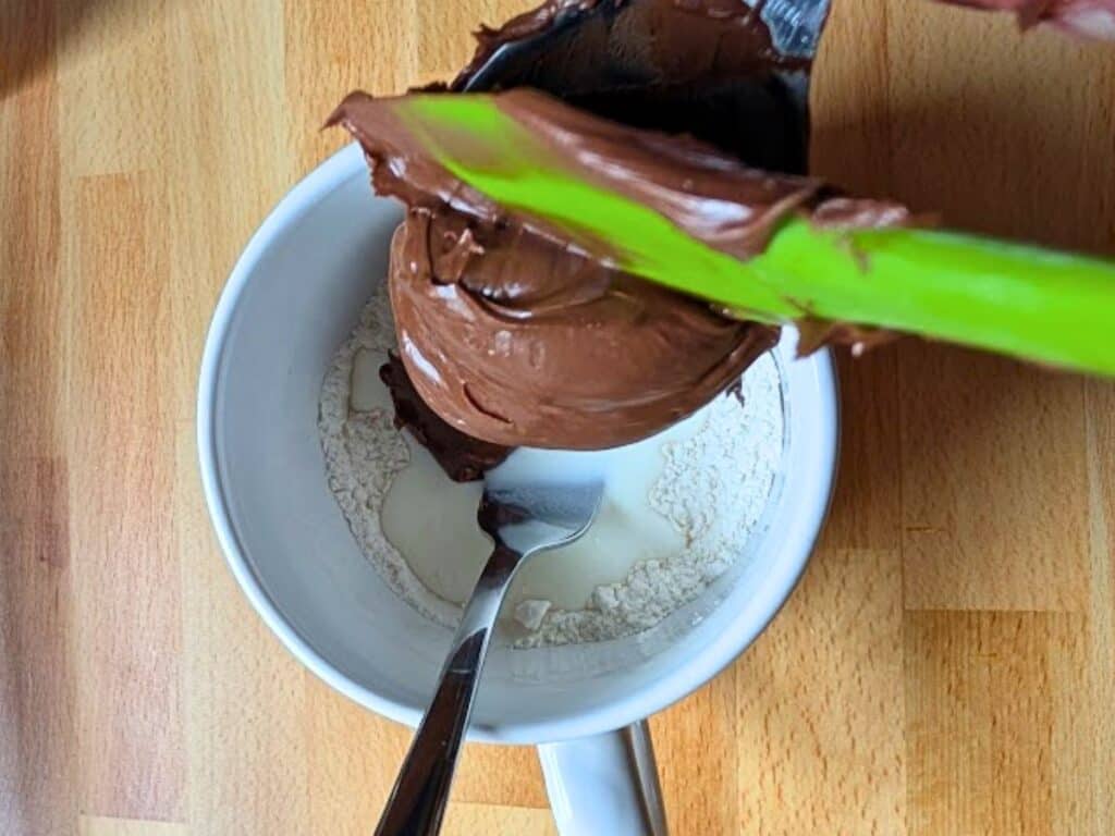 A green spatula adds chocolate spread to a white mug containing flour and a small amount of liquid, with a spoon inside the mug.