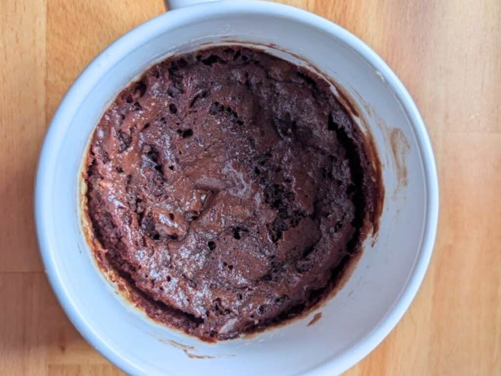A chocolate mug cake sits in a white mug on a wooden surface, showing a moist, slightly cracked top.