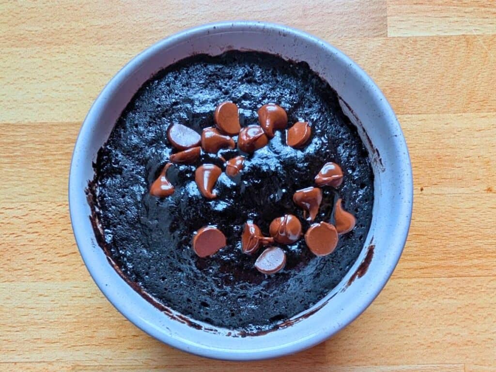 A white bowl filled with chocolate mug cake topped with melted chocolate chips sits on a wooden surface.