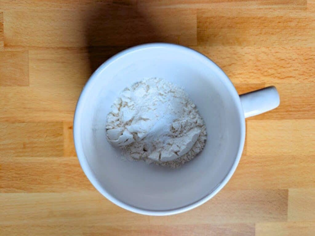 A white mug containing a small amount of white powder, likely flour, is placed on a wooden surface.
