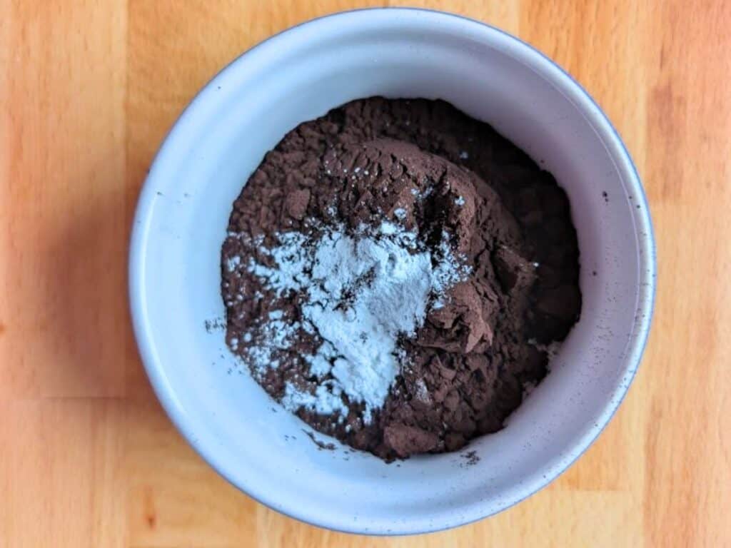 A white bowl containing cocoa powder and baking powder on a wooden surface.