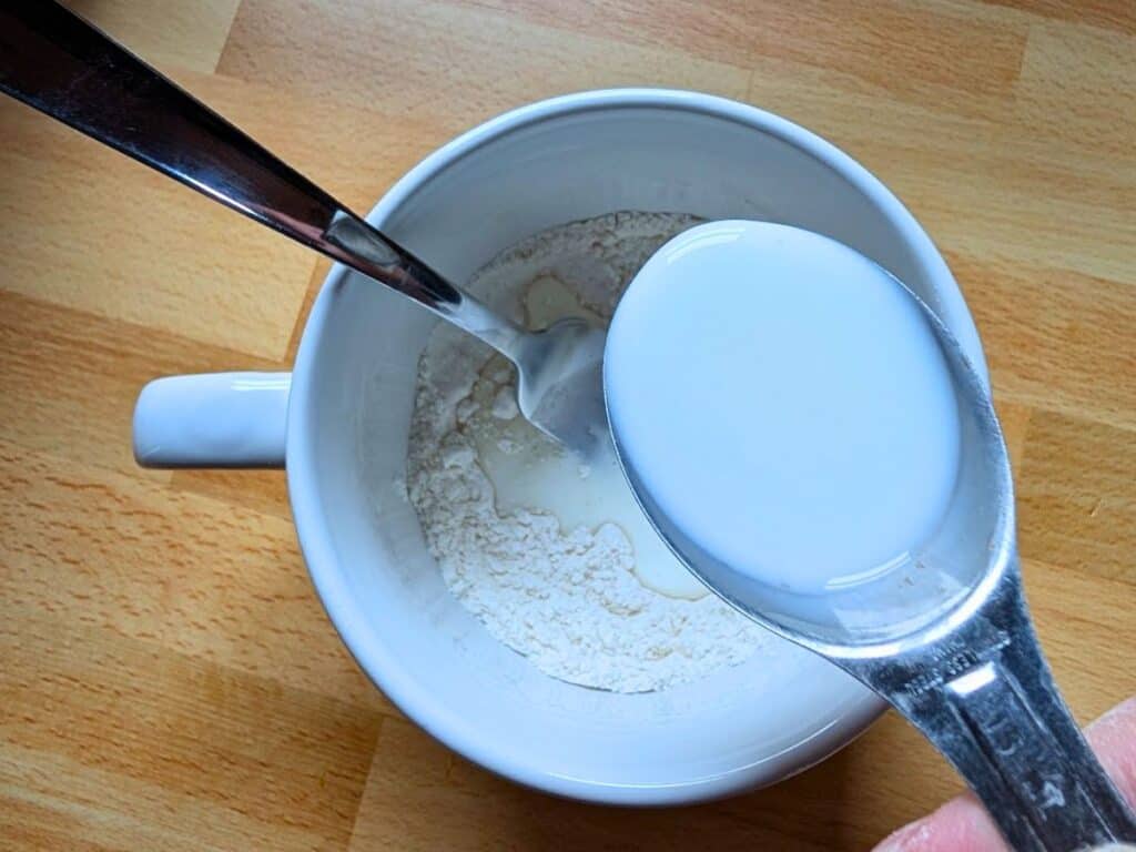 A spoon holds liquid over a white mug containing flour and a spoon, set on a wooden surface.