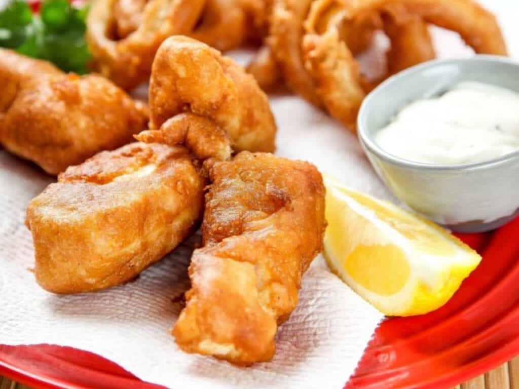 Battered and fried fish pieces with onion rings, a lemon wedge, and a small bowl of white dipping sauce on a red plate.