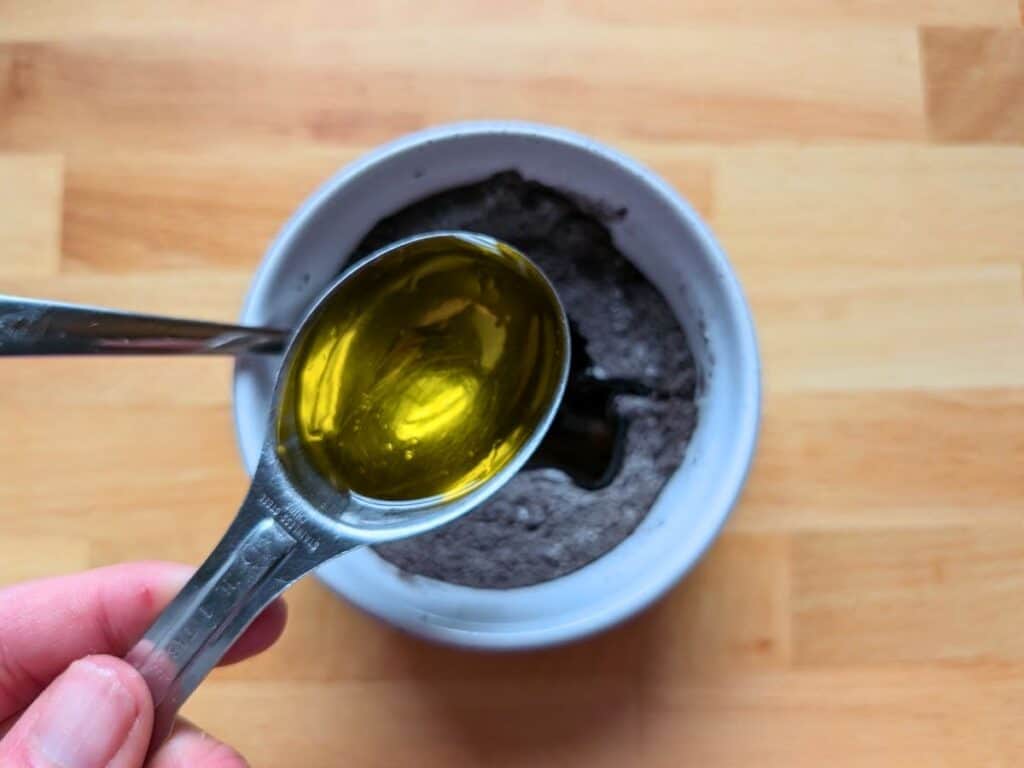 A hand holds a measuring spoon filled with olive oil above a white bowl containing a gray powder mixture on a wooden surface.