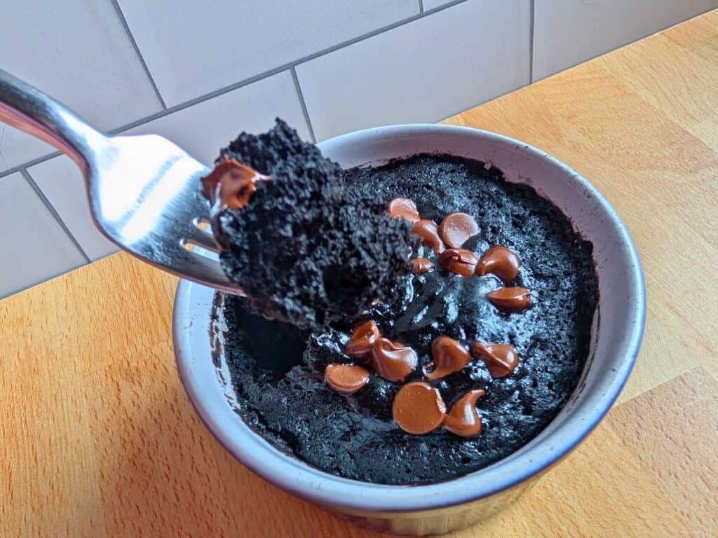 A fork holding a portion of chocolate mug cake with chocolate chips, above a ceramic mug on a wooden surface.