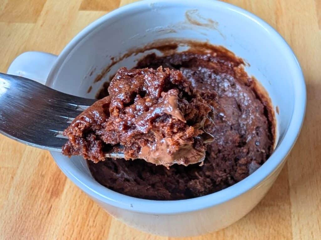 A close-up of a chocolate mug cake in a white mug, with a fork lifting a portion of the cake.