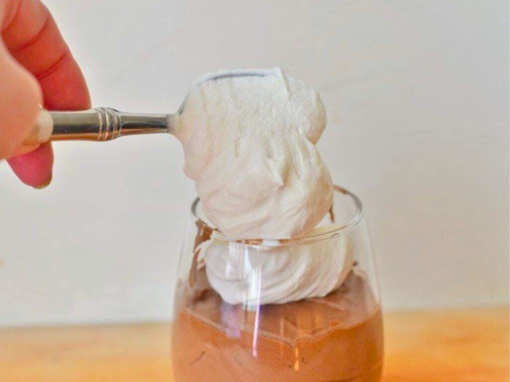 A hand holds a spoon with whipped cream, placing it on top of chocolate mousse in a glass.