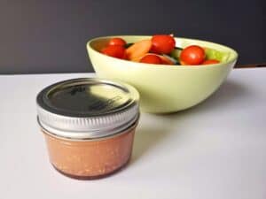 A small sealed glass jar of homemade dijon vinaigrette sits in front of a light green bowl filled with lettuce, cherry tomatoes, and sliced carrots on a white surface.