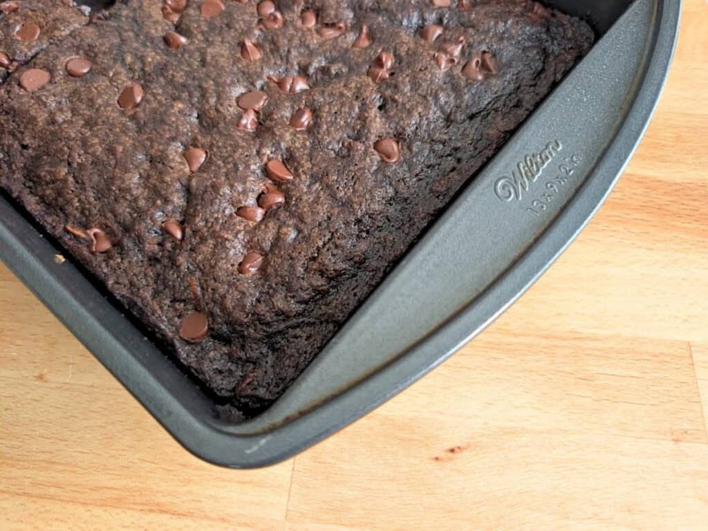 A metal pan with baked chocolate brownies topped with chocolate chips sits on a wooden surface, with one corner piece missing.