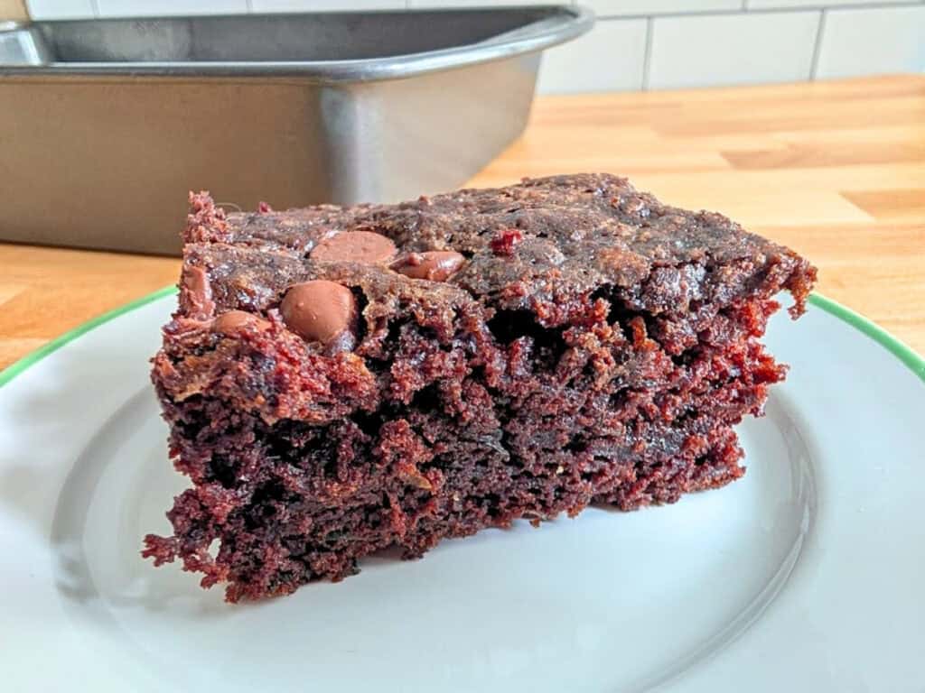 A slice of chocolate cake with chocolate chips on a white plate, with a metal baking pan and wooden countertop in the background.