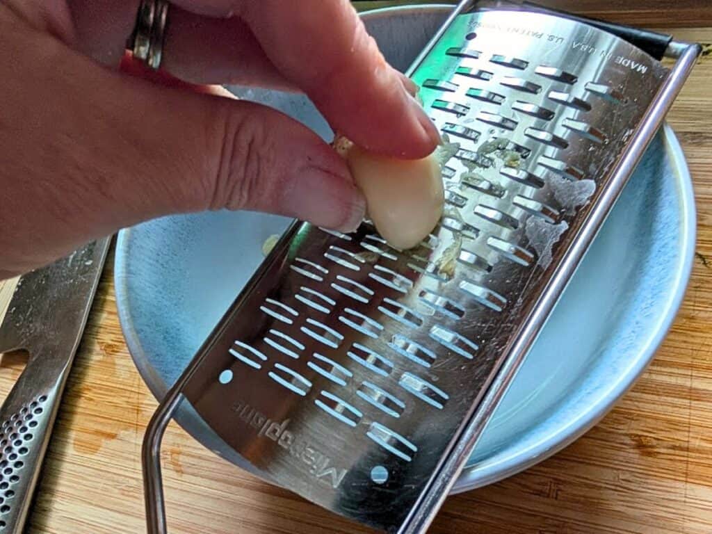 A hand grates a garlic clove over a metal grater positioned above a white bowl on a wooden surface.
