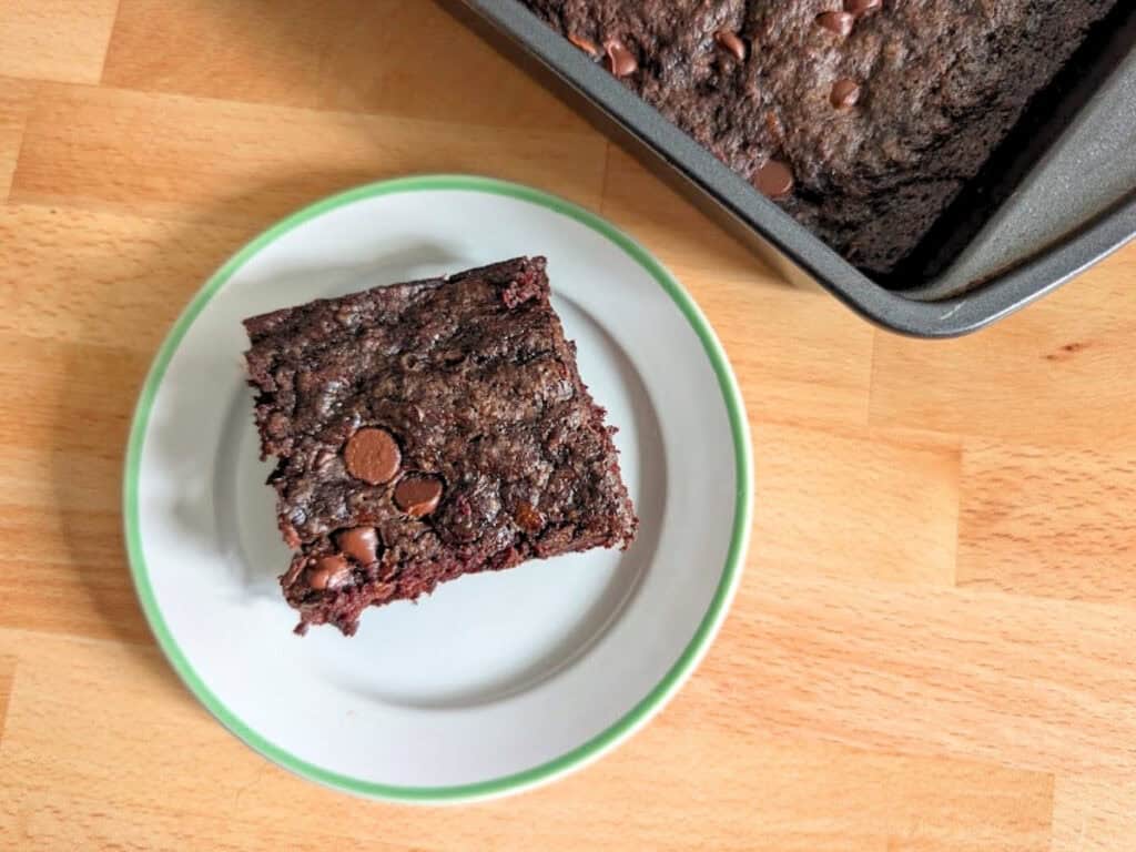 A square piece of chocolate brownie with chocolate chips sits on a white plate next to a pan of brownies on a wooden surface.