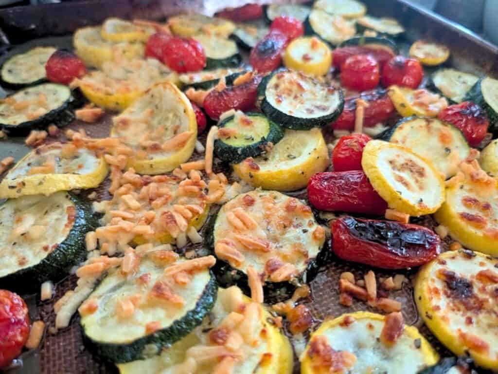 Roasted zucchini, yellow squash, and grape tomatoes topped with melted shredded cheese on a baking sheet.