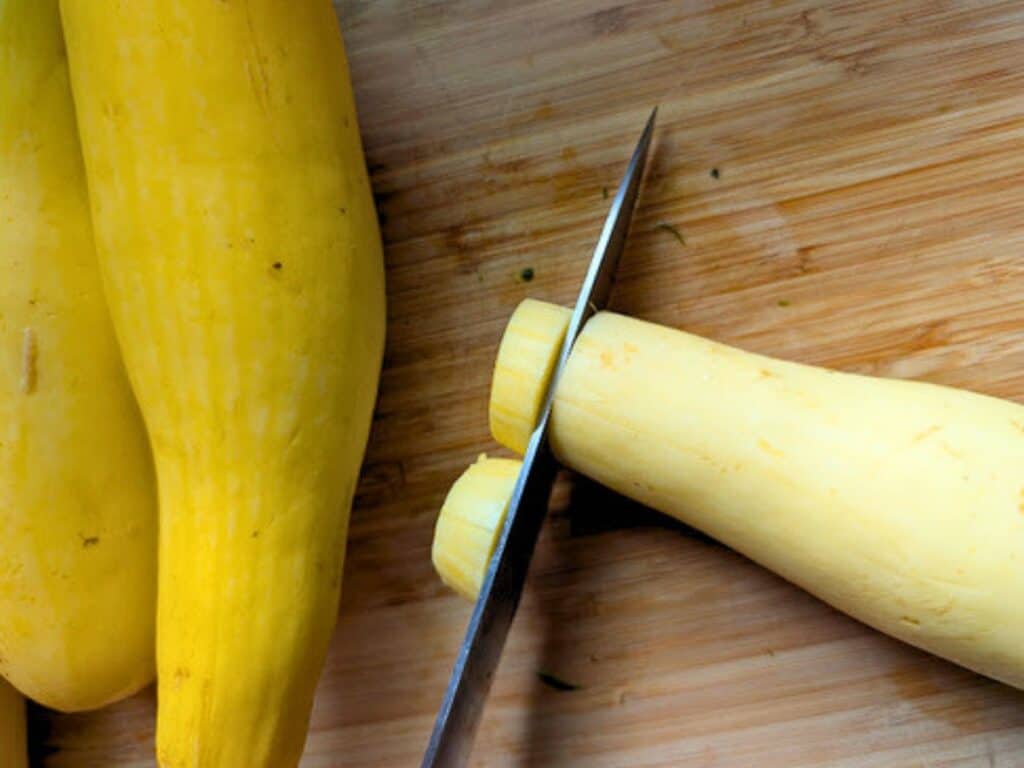 A knife slices the end off a yellow squash on a wooden cutting board, with more squash lying beside it.