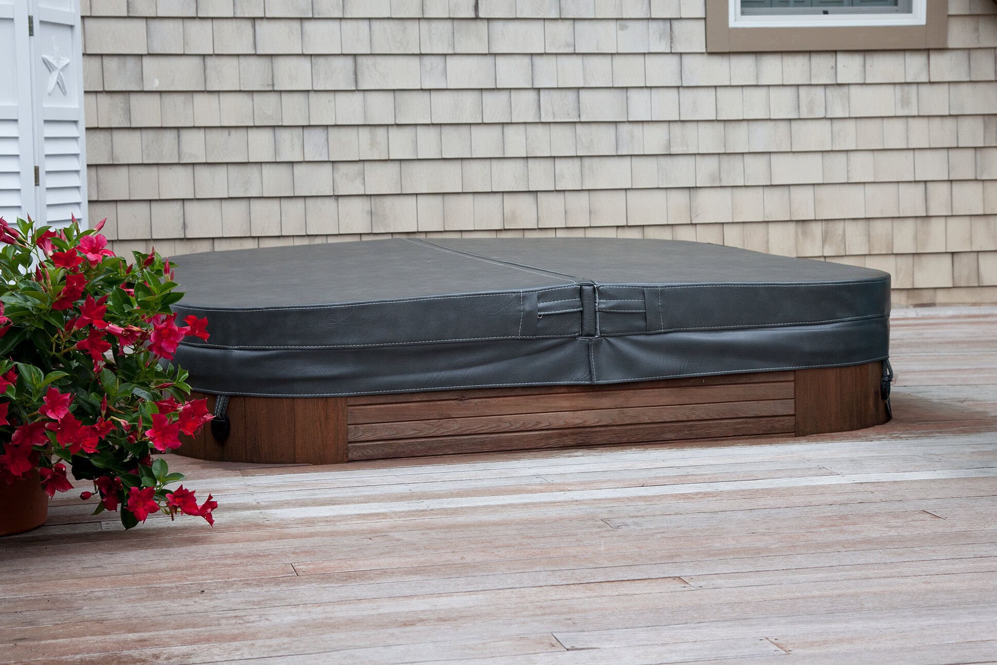 Black hot tub cover on a wooden spa beside red flowering plants, showcasing outdoor spa protection.