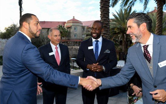 Professional businessmen shaking hands during Houston EB5 investment event in an outdoor setting with palm trees and hotel background.