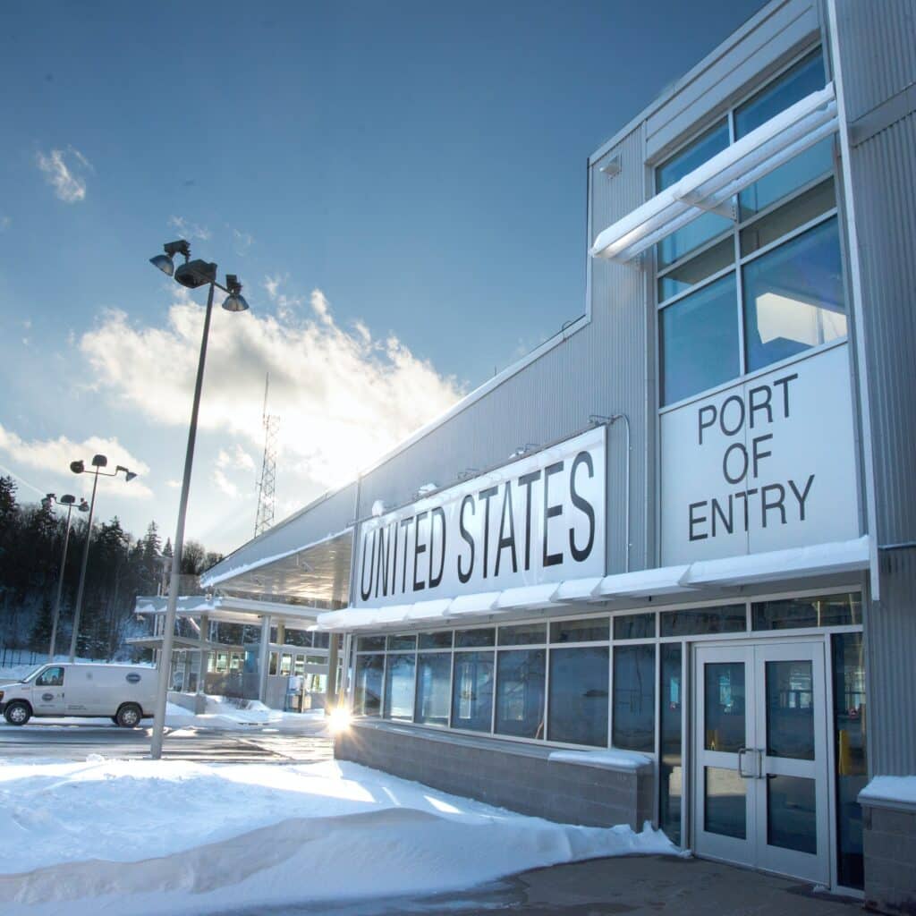 Snow-covered US port of entry building with "UNITED STATES" and "PORT OF ENTRY" signage under a bright blue sky.