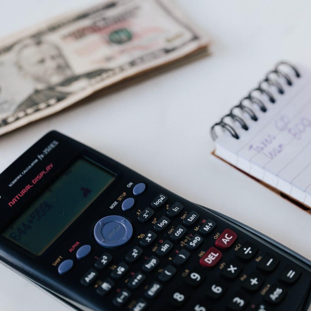 $20 bills placed on a white surface, with a calculator, a notepad, and a person writing notes, illustrating investment calculations for Houston EB5 visa programs.