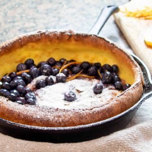 Baked Dutch Baby pancake in a cast iron pan with blueberries, icing sugar and orange zest. Sliced oranges in the background on a wooden cutting board.